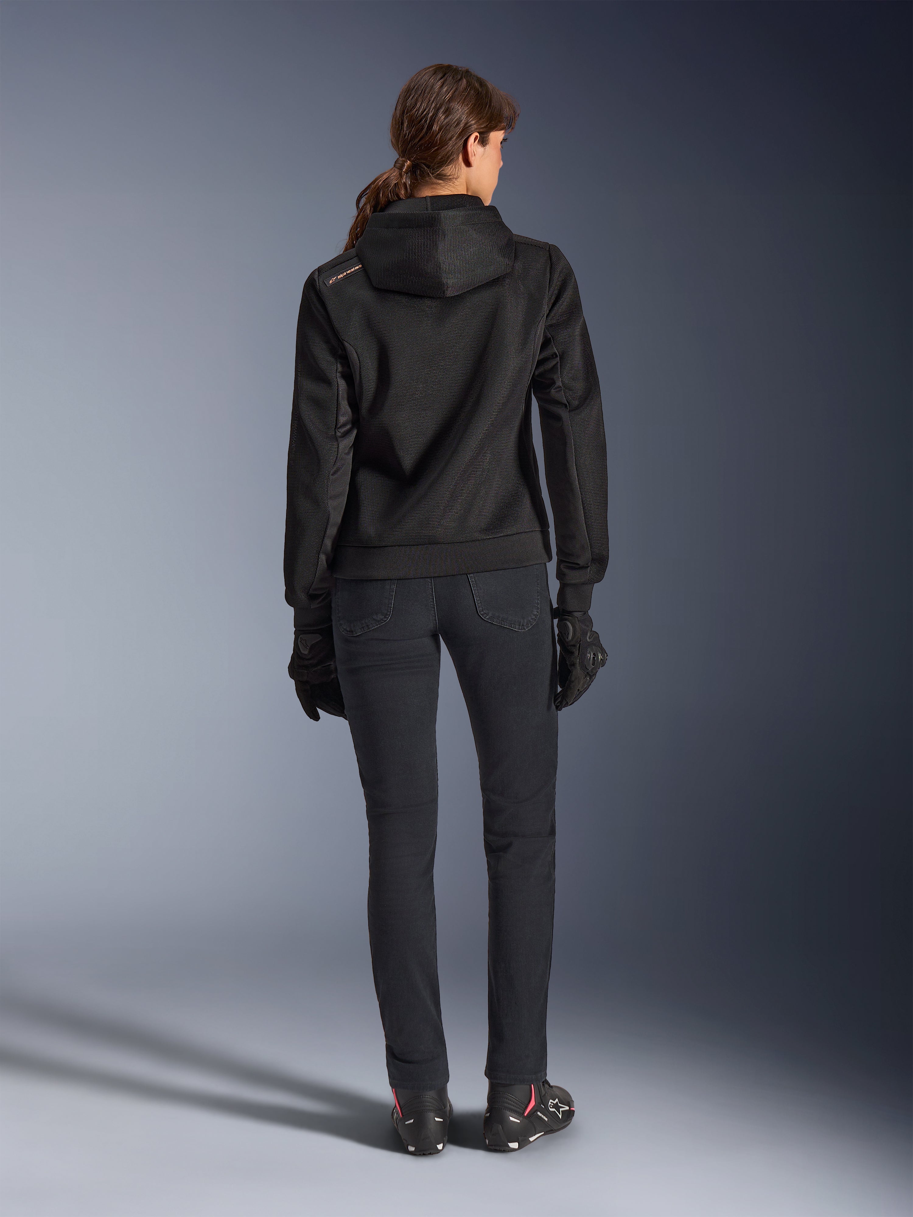 Alpinestars Stella Chrome Superairflow Hoodie, sweat à capuche technique de moto pour femme, maille 3D noire avec petit logo orange sur l'épaule, vue de dos, porté avec un pantalon de moto noir, des gants et des bottes noirs, conçu pour la conduite sportive urbaine
