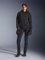 Modèle féminin portant le sweat à capuche Alpinestars Stella Chrome Superairflow noir, associé à un pantalon de moto technique et des gants noirs, doté d'une construction en maille 3D et d'un logo cuivré sur la poitrine, vêtement de moto sport urbain pour femme
