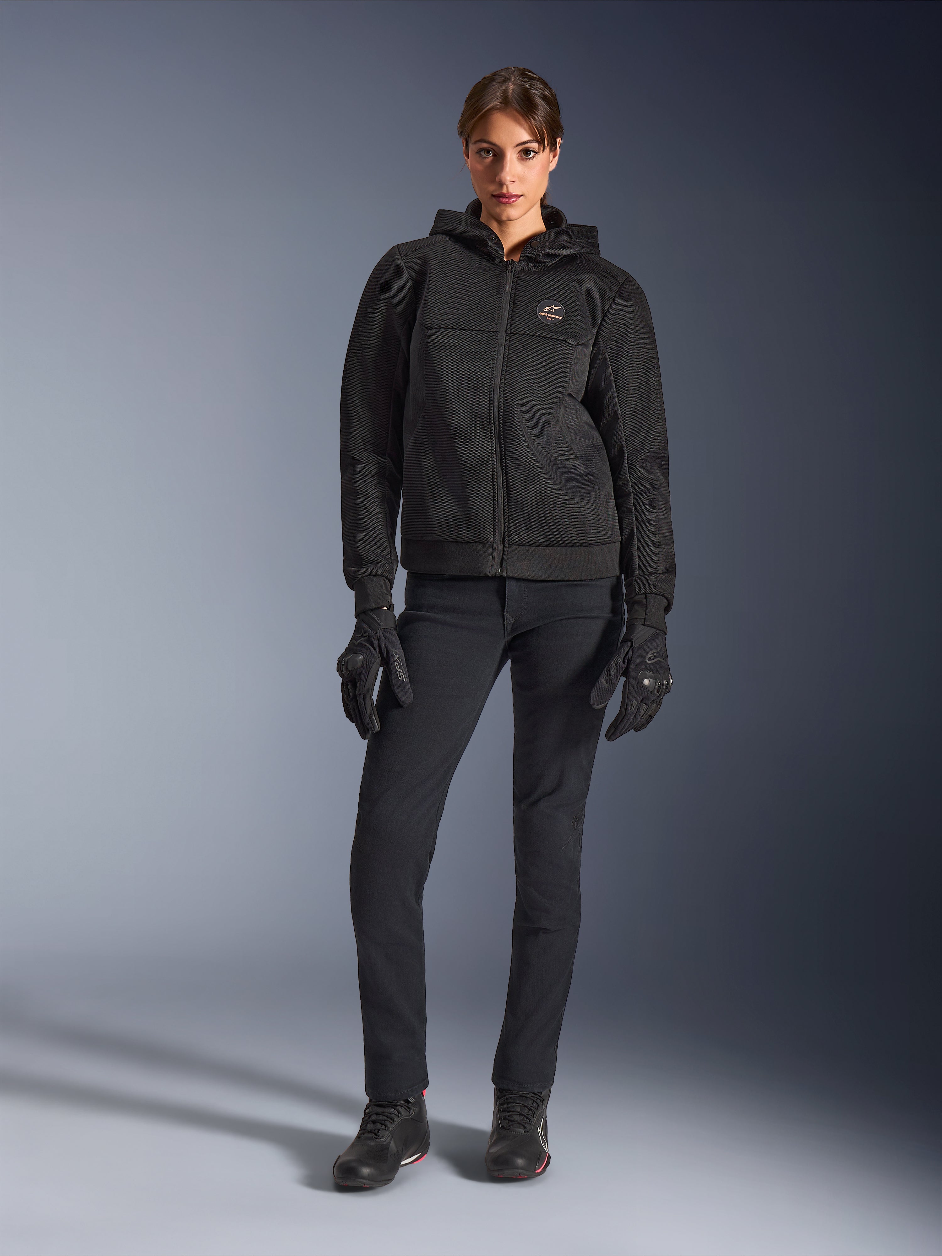 Modèle féminin portant le sweat à capuche Alpinestars Stella Chrome Superairflow noir, associé à un pantalon de moto technique et des gants noirs, doté d'une construction en maille 3D et d'un logo cuivré sur la poitrine, vêtement de moto sport urbain pour femme