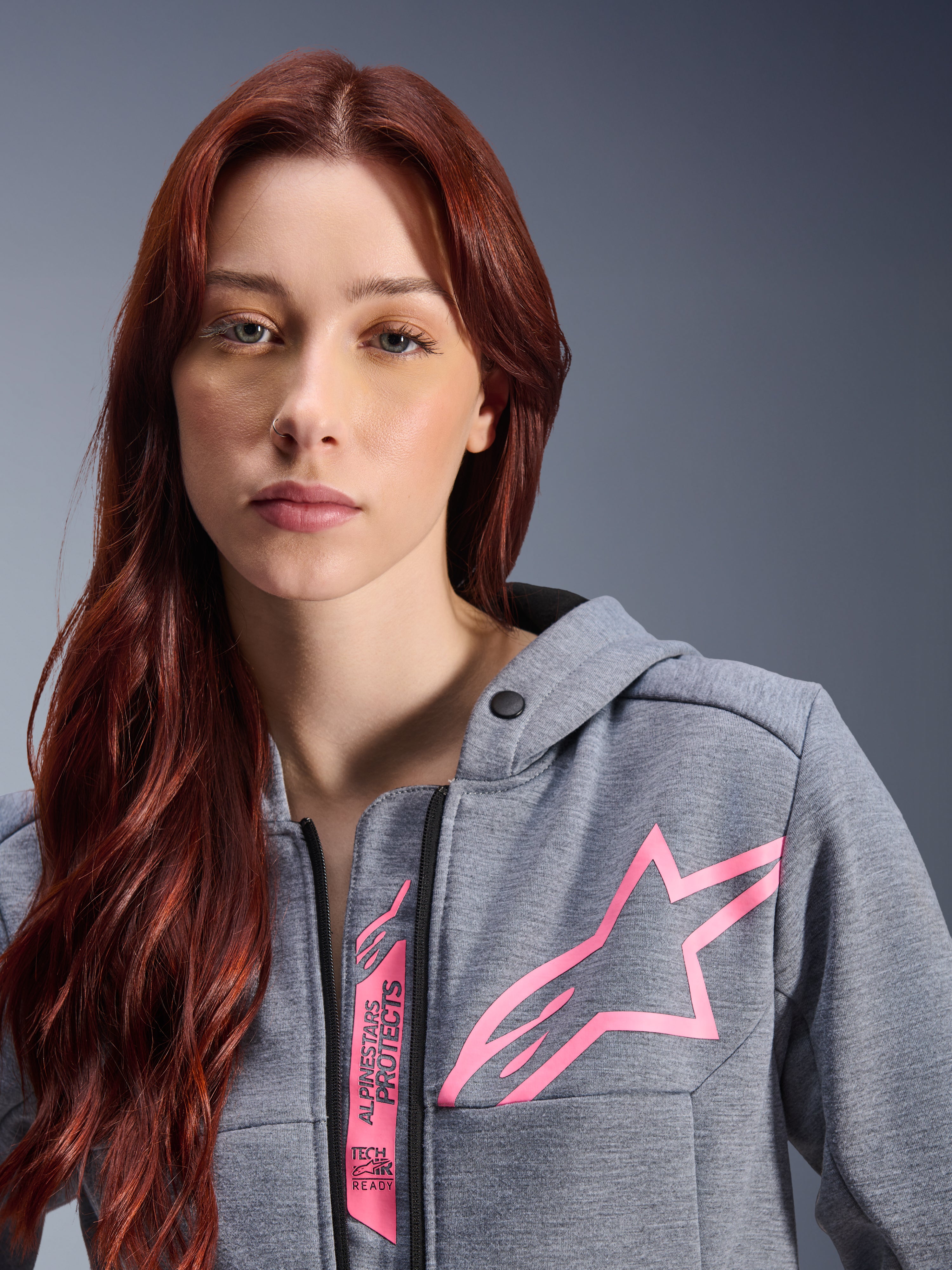 Woman wearing Alpinestars Stella Chrome V2 Sport Hoodie, Gray Heather Pink Fluo, close-up shot showing the pink Alpinestars star logo and Tech-Air Ready branding, urban motorcycle apparel.