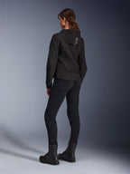 Alpinestars Stella Radium Tech Hoodie, sweat à capuche de moto urbain noir pour femme, vue arrière montrant le logo 'ALPIN' sur la capuche, porté avec un pantalon de moto technique noir et des bottes noires, matière technique légère et coupe-vent.