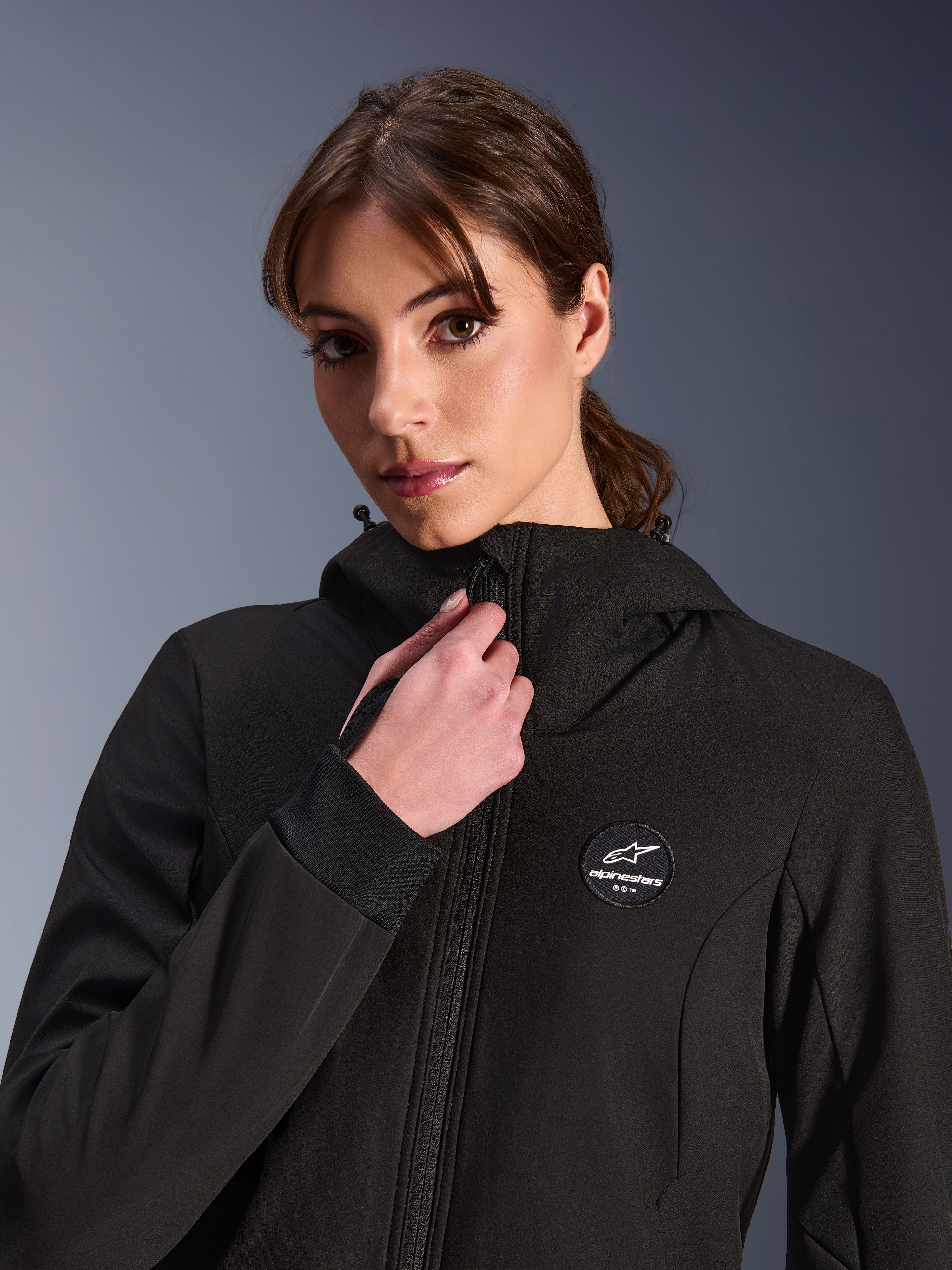 Hoodie Stella Radium Tech Femme, noir, porté par une femme ajustant la fermeture éclair frontale, avec écusson logo Alpinestars circulaire sur la poitrine, poignets côtelés extensibles et construction textile coupe-vent pour la conduite urbaine