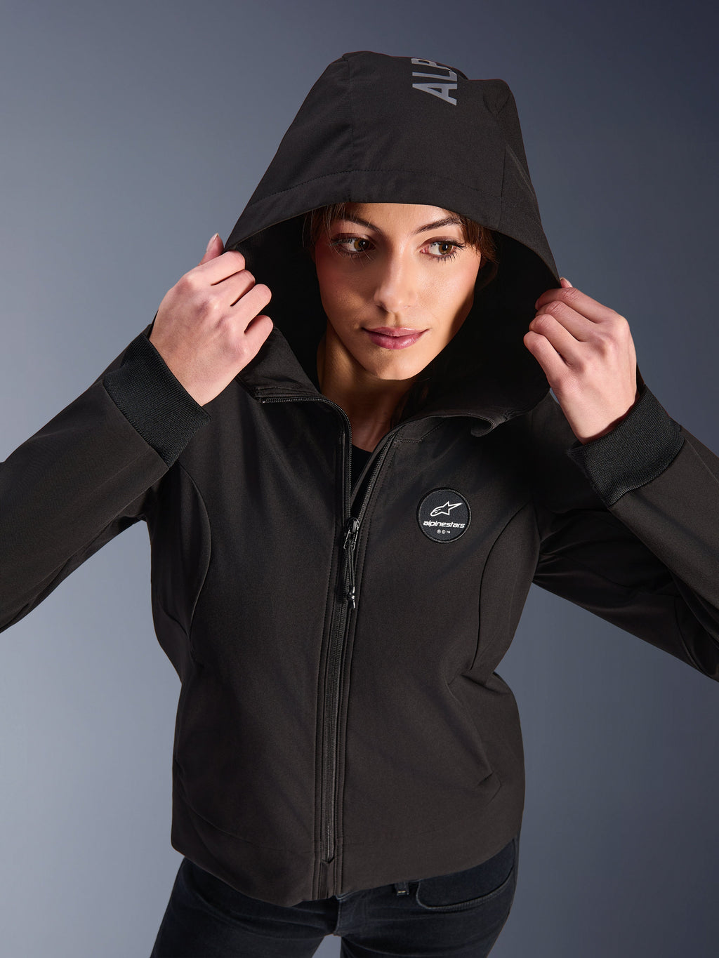Femme portant le sweat à capuche Alpinestars Stella Radium Tech en noir, hoodie de moto urbain avec la capuche relevée, présentant un logo Alpinestars circulaire sur la poitrine, des poignets côtelés et une construction coupe-vent à 3 couches
