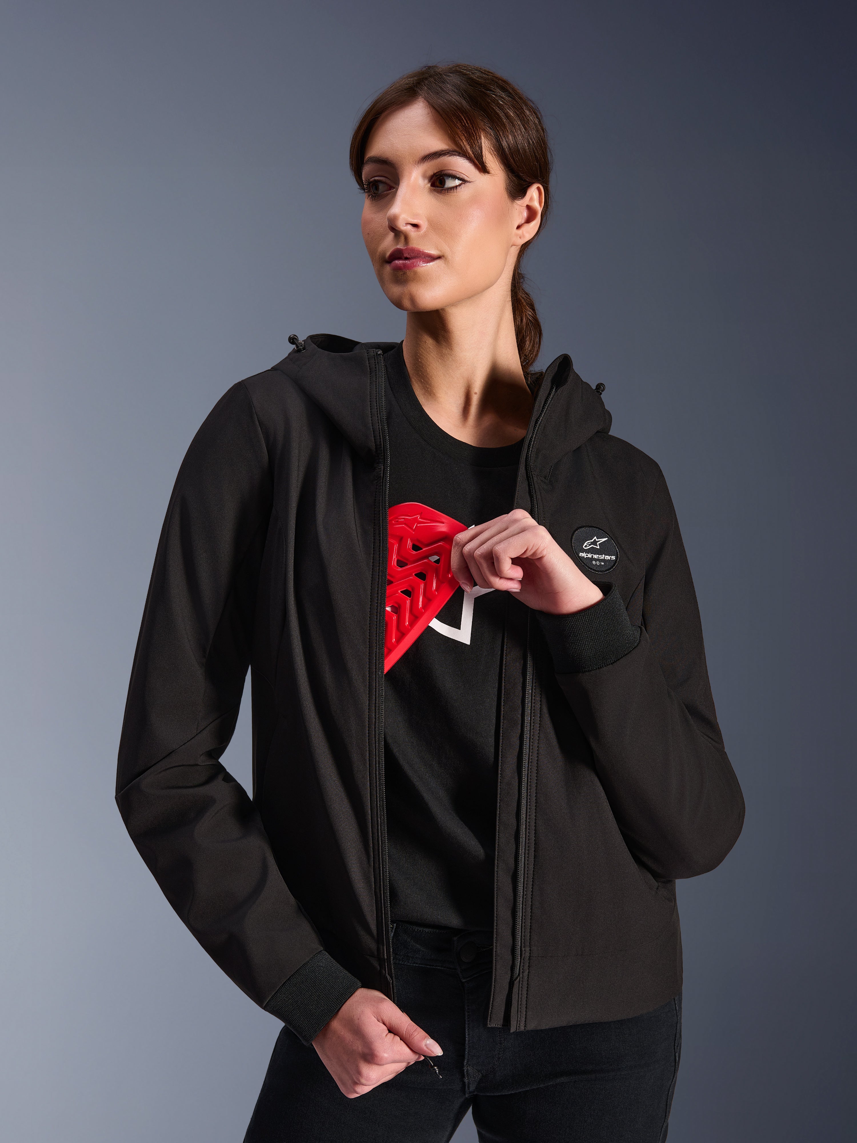 Alpinestars Stella Radium Tech Hoodie pour femme, sweat à capuche de moto urbain noir, mannequin démontrant la protection Nucleon Flex Plus rouge à l'intérieur, coque technique légère à 3 couches coupe-vent avec poignets côtelés et écusson logo, vêtement de protection moto