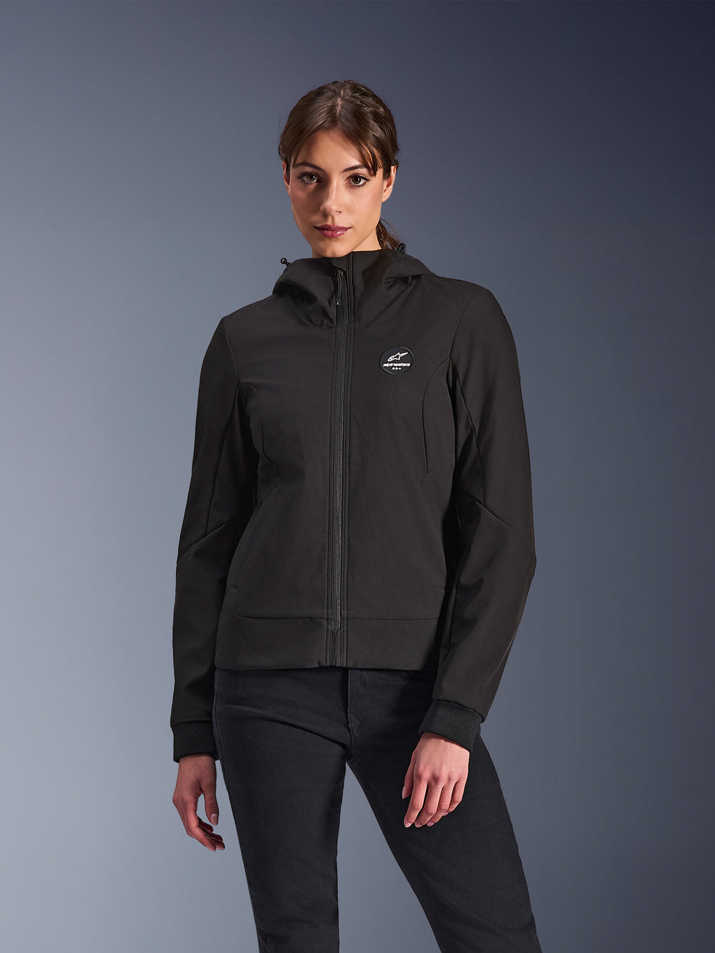 Alpinestars Stella Radium Tech Hoodie, veste de moto à capuche pour femme, noire, design zippé avec logo circulaire sur la poitrine et capuche, vêtement technique de conduite urbaine, porté par un mannequin