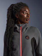 Modèle féminin portant le sweat à capuche Alpinestars Stella Radium Tech Hoodie en gris foncé et noir, avec une fermeture éclair corail vif, un logo circulaire sur la poitrine et une coque coupe-vent à col haut, tenue de moto urbaine décontractée pour femmes