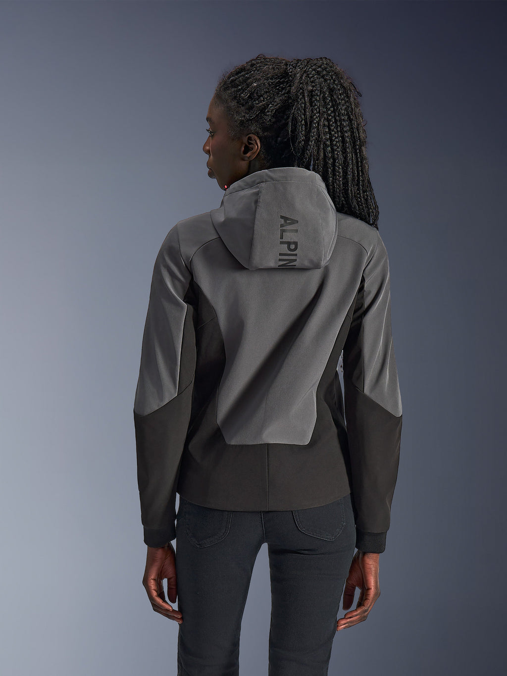 Femme portant le sweat à capuche Alpinestars Stella Radium Tech, vue de dos, Deep Gray Black, coque bicolore grise et noire à 3 couches coupe-vent, logo Alpinestars sur la capuche, vêtement de moto urbain à coupe athlétique