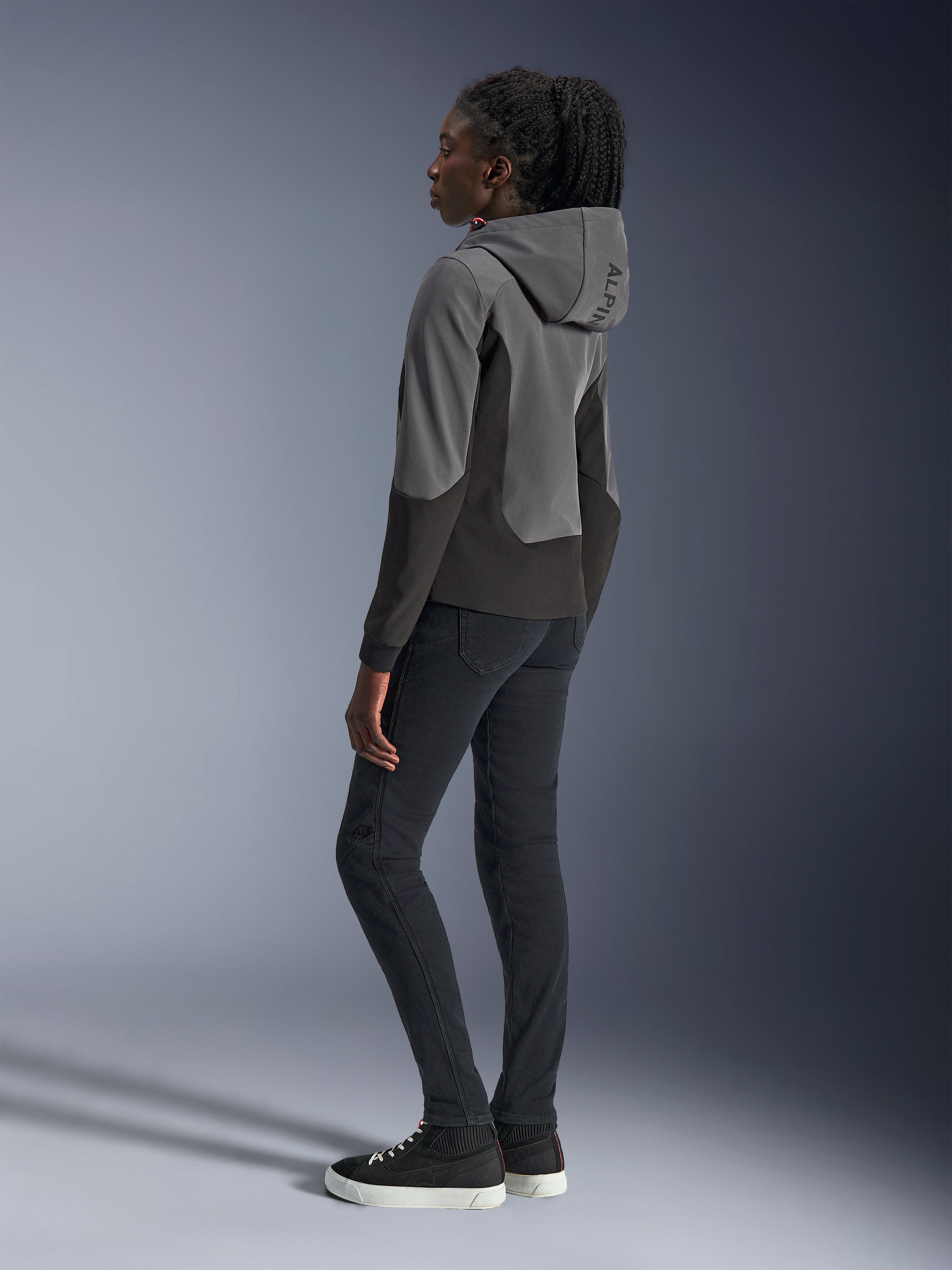 Alpinestars Stella Radium Tech Hoodie pour femme, gris foncé et noir, équipement de moto urbain, vue de dos montrant la capuche avec marquage Alpinestars, porté avec un pantalon slim noir et des baskets montantes