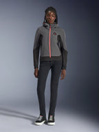 Modèle féminin portant le sweat à capuche Alpinestars Stella Radium Tech, gris foncé et noir avec une fermeture éclair orange vif, sweat à capuche de moto urbain, associé à un pantalon slim noir et des baskets, tenue de performance athlétique décontractée pour femme.