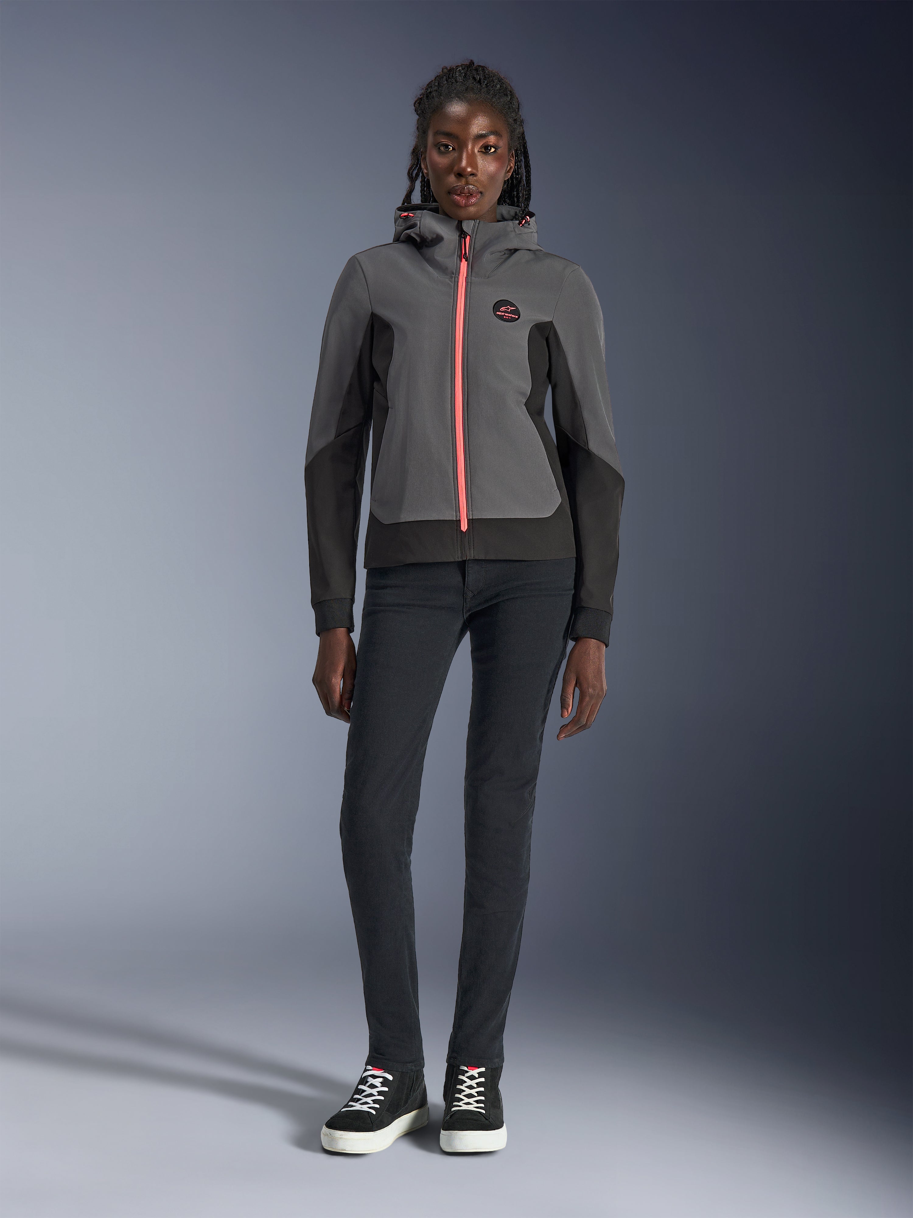 Modèle féminin portant le sweat à capuche Alpinestars Stella Radium Tech, gris foncé et noir avec une fermeture éclair orange vif, sweat à capuche de moto urbain, associé à un pantalon slim noir et des baskets, tenue de performance athlétique décontractée pour femme.