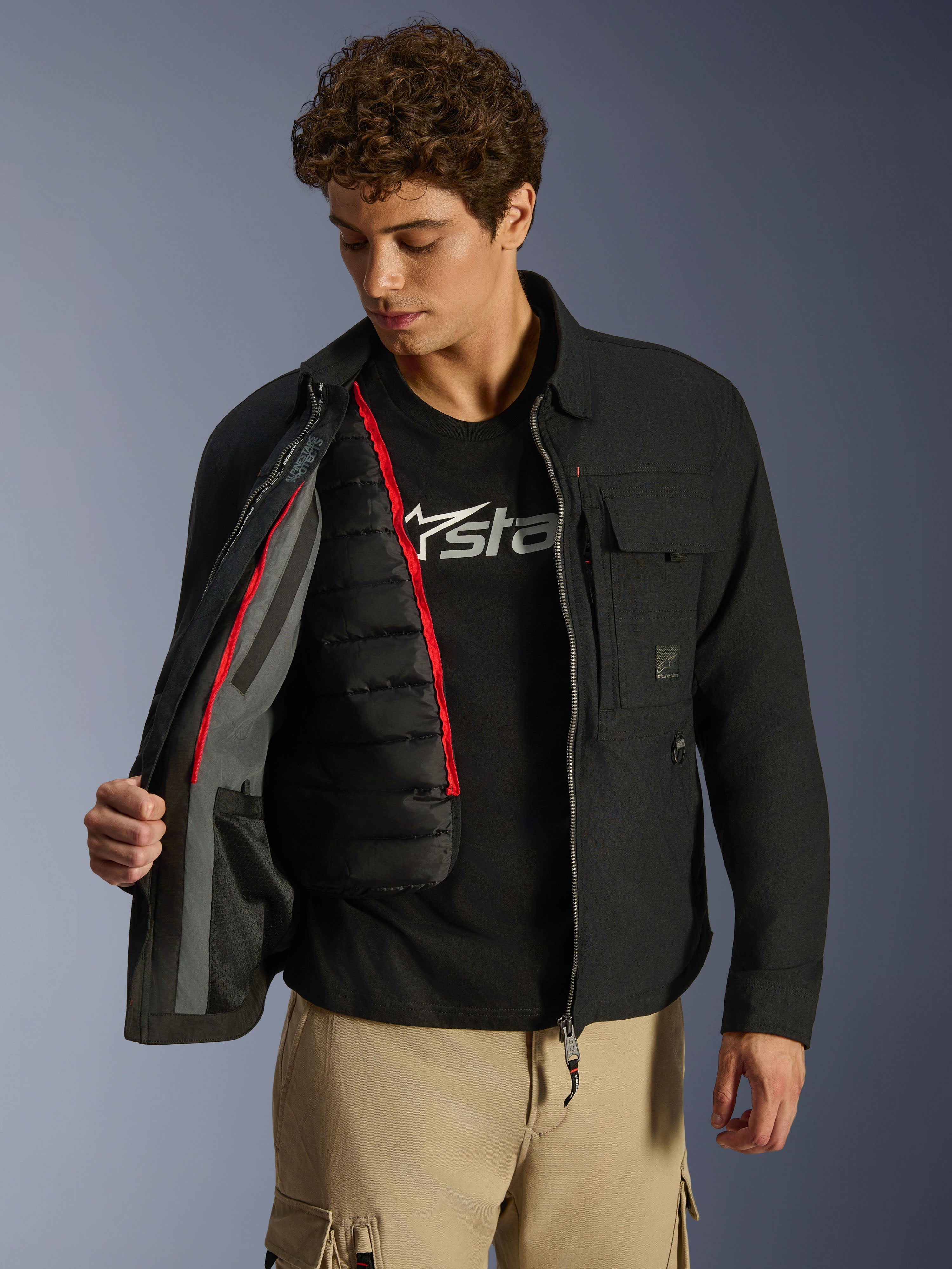 Surchemise Alpinestars Bryon WR, noire, veste de moto urbaine présentée ouverte pour révéler un gilet thermique matelassé noir amovible avec liseré rouge, portée avec un pantalon cargo beige et un t-shirt à logo, design fonctionnel d'inspiration workwear