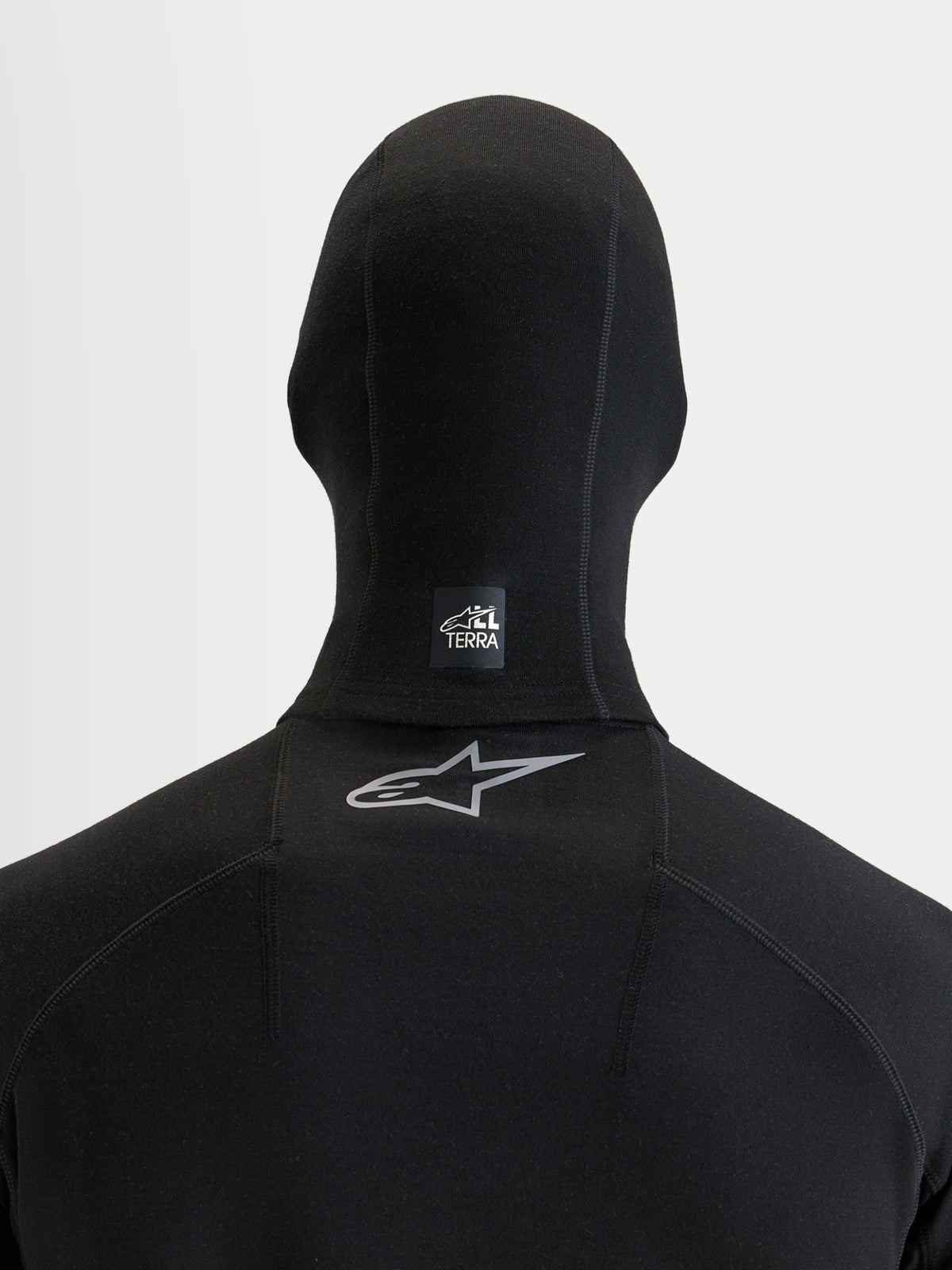 Alpinestars At 8 Merino Cagoule, cagoule de moto noire, vue de dos montrant une étiquette All Terra sur le cou et un logo Astar réfléchissant sur le haut du dos, construction en mélange de laine mérinos thermique