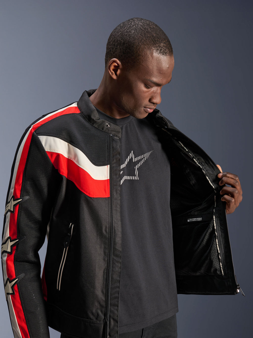 Veste Alpinestars T-Dyno Air, noir, rouge vif et écru, mannequin homme ouvrant la veste pour montrer la doublure interne et le compartiment imperméable, veste textile d'inspiration vintage avec panneaux en maille 3D et logos étoiles, équipement de moto moderne