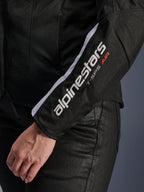 Veste Alpinestars Stella T-SPS Air V2, veste de moto pour femme, noir et blanc, gros plan de la manche avec logo Alpinestars blanc et texte T-SPS AIR, construction en mesh et textile pour la circulation de l'air, portée avec un pantalon de moto noir
