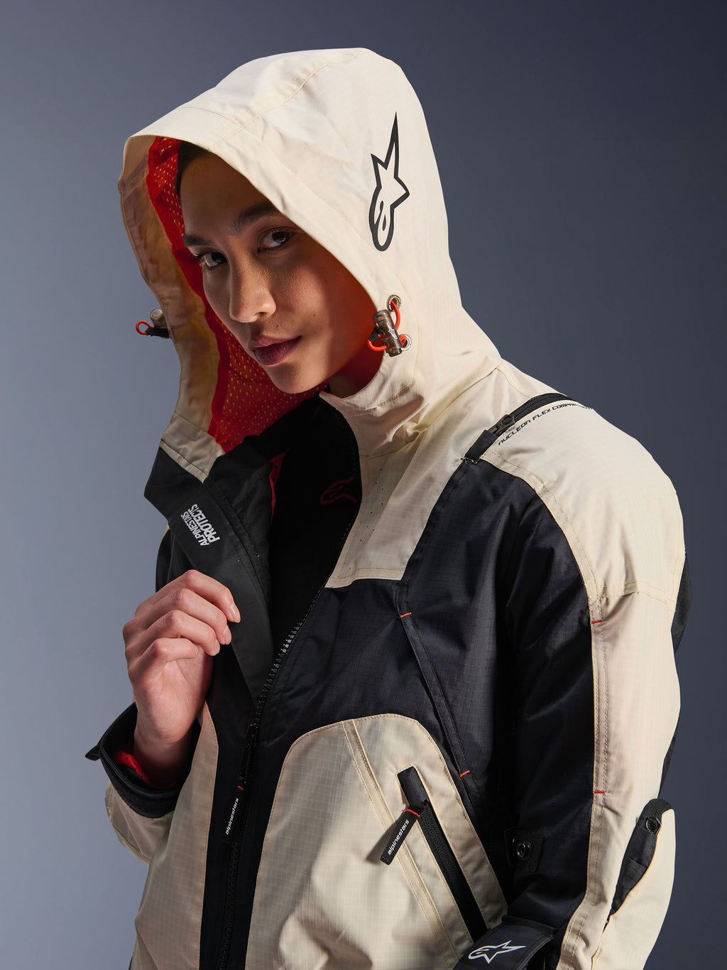 Veste Alpinestars Molly Air en Black Almond, portée par un mannequin femme avec la capuche relevée, présentant un design color-block crème et noir, une doublure en mesh orange vif et des éléments de design urbain technique incluant un logo étoile et des détails de zips.