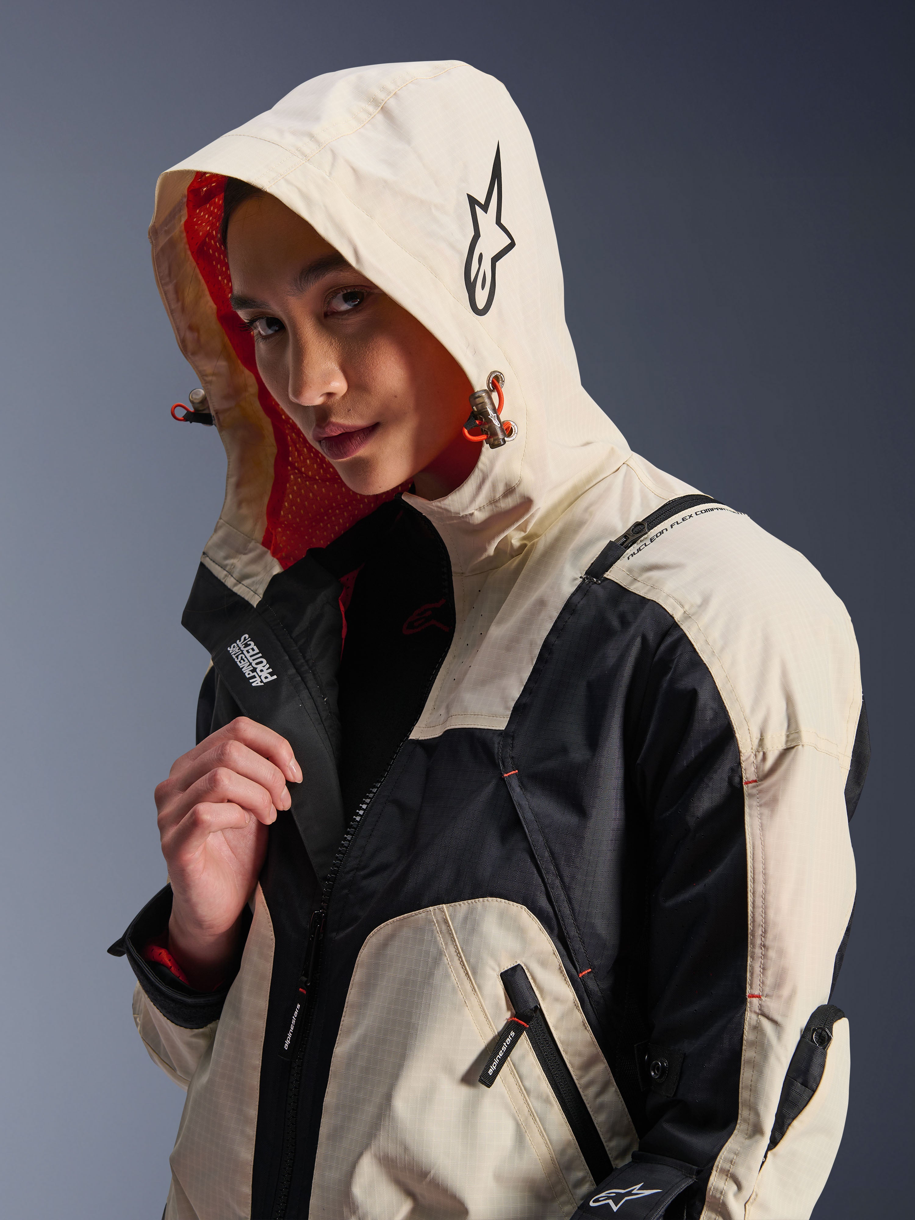 Veste Alpinestars Molly Air en Black Almond, portée par un mannequin femme avec la capuche relevée, présentant un design color-block crème et noir, une doublure en mesh orange vif et des éléments de design urbain technique incluant un logo étoile et des détails de zips.