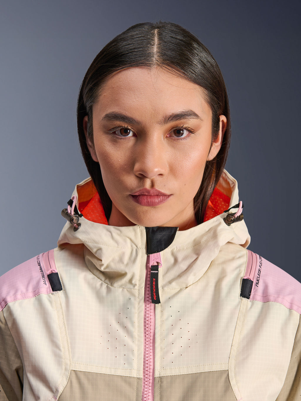 Gros plan d'un mannequin femme portant la veste Alpinestars Molly Air en Sand Pink Almond, avec col haut et capuche à doublure orange vif, fermeture éclair rose, tissu ripstop et poitrine perforée au laser pour la ventilation, équipement de protection moto urbain