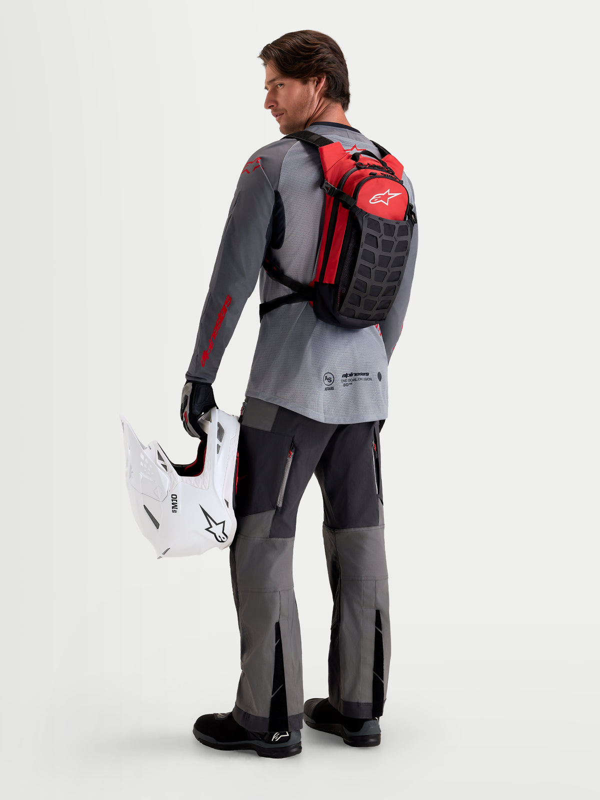 Alpinestars Sac À Dos AT-9, sac à dos de motocross noir et rouge vif, porté par un modèle de dos tenant un casque blanc, construction textile légère pour le circuit et le paddock