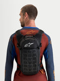 Sac à dos Alpinestars AT-9, sac à dos de motocross porté par un modèle, construction en textile noir avec logo Alpinestars blanc, design compact pour le circuit et l'usage quotidien