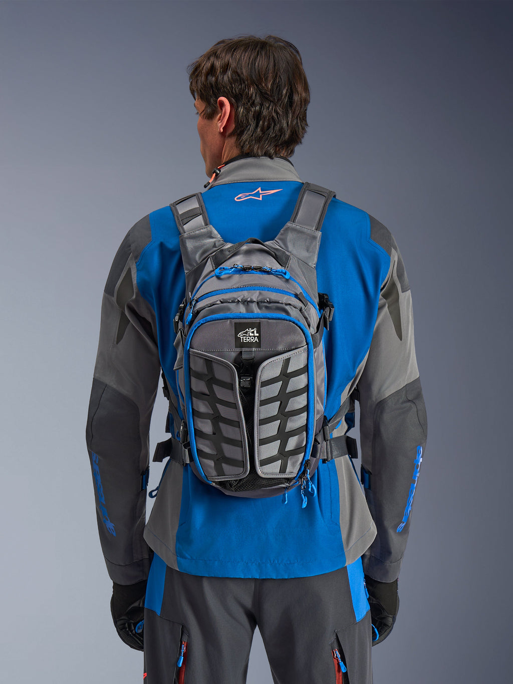 Sac à dos Alpinestars AT-15, sac à dos de moto aventure, gris foncé et gris profond, porté par un motard sur une veste bleue et grise, doté du marquage All Terra, d'un design structurel ergonomique, de sangles réglables et de touches de bleu sur les fermetures éclair.