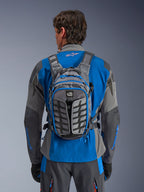 Sac à dos Alpinestars AT-15, sac à dos de moto aventure, gris foncé et gris profond, porté par un motard sur une veste bleue et grise, doté du marquage All Terra, d'un design structurel ergonomique, de sangles réglables et de touches de bleu sur les fermetures éclair.