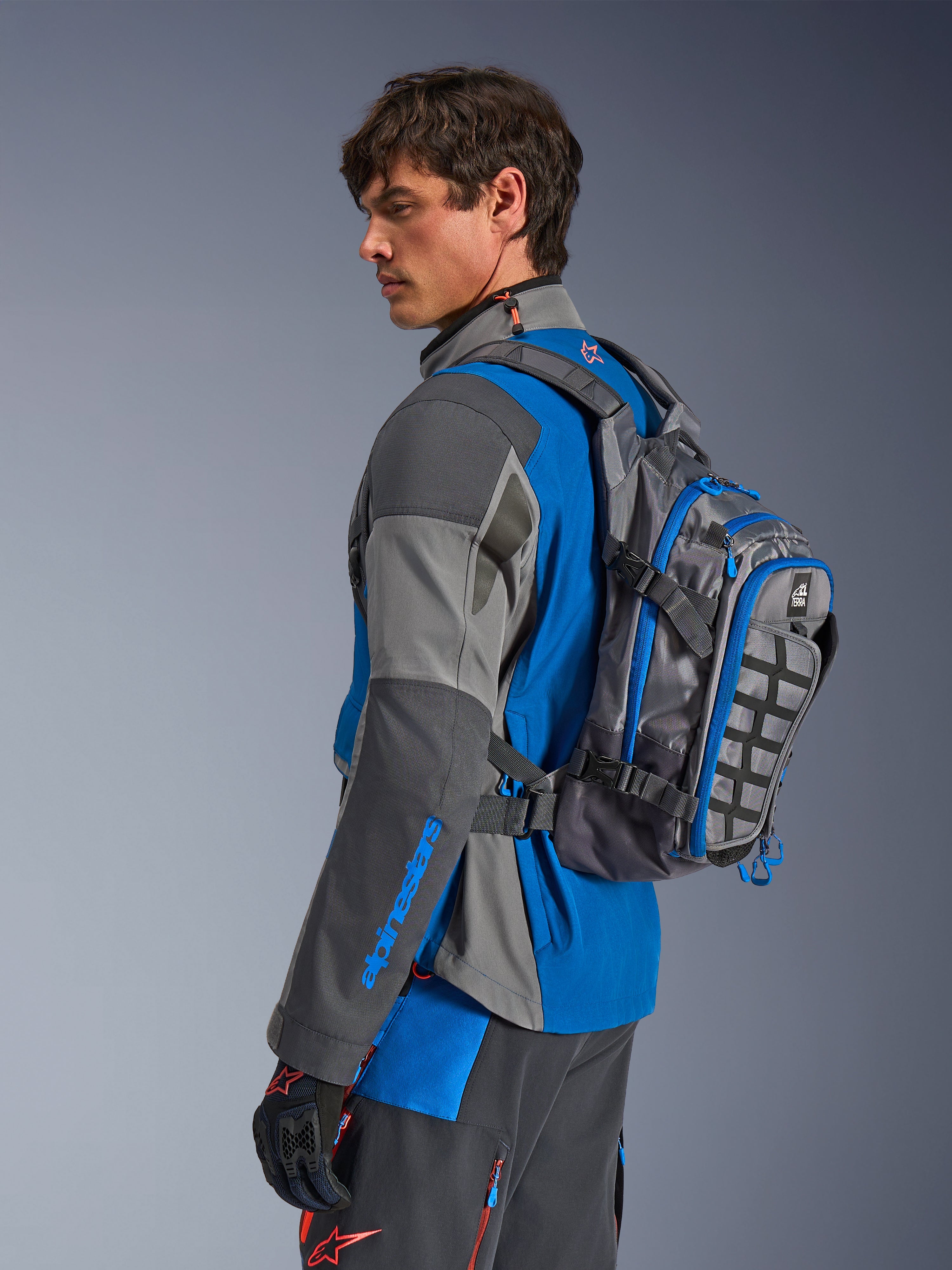 Sac à dos Alpinestars AT-15, sac à dos d'aventure moto, gris foncé avec des accents bleus, porté par un motard en tenue technique coordonnée grise et bleue, doté d'un panneau MOLLE et d'une construction robuste pour l'aventure tout-terrain.