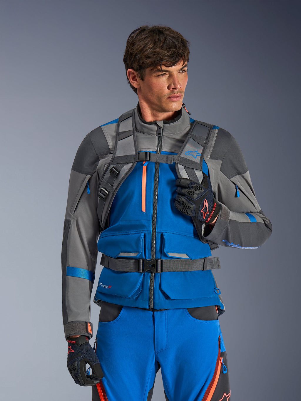 Sac à dos Alpinestars AT-15, sac à dos de moto, gris foncé, porté par un pilote avec une veste et un pantalon d'aventure bleus et gris, doté de bretelles techniques grises avec boucle de poitrine magnétique et sangle ventrale, conception robuste pour la conduite tout-terrain