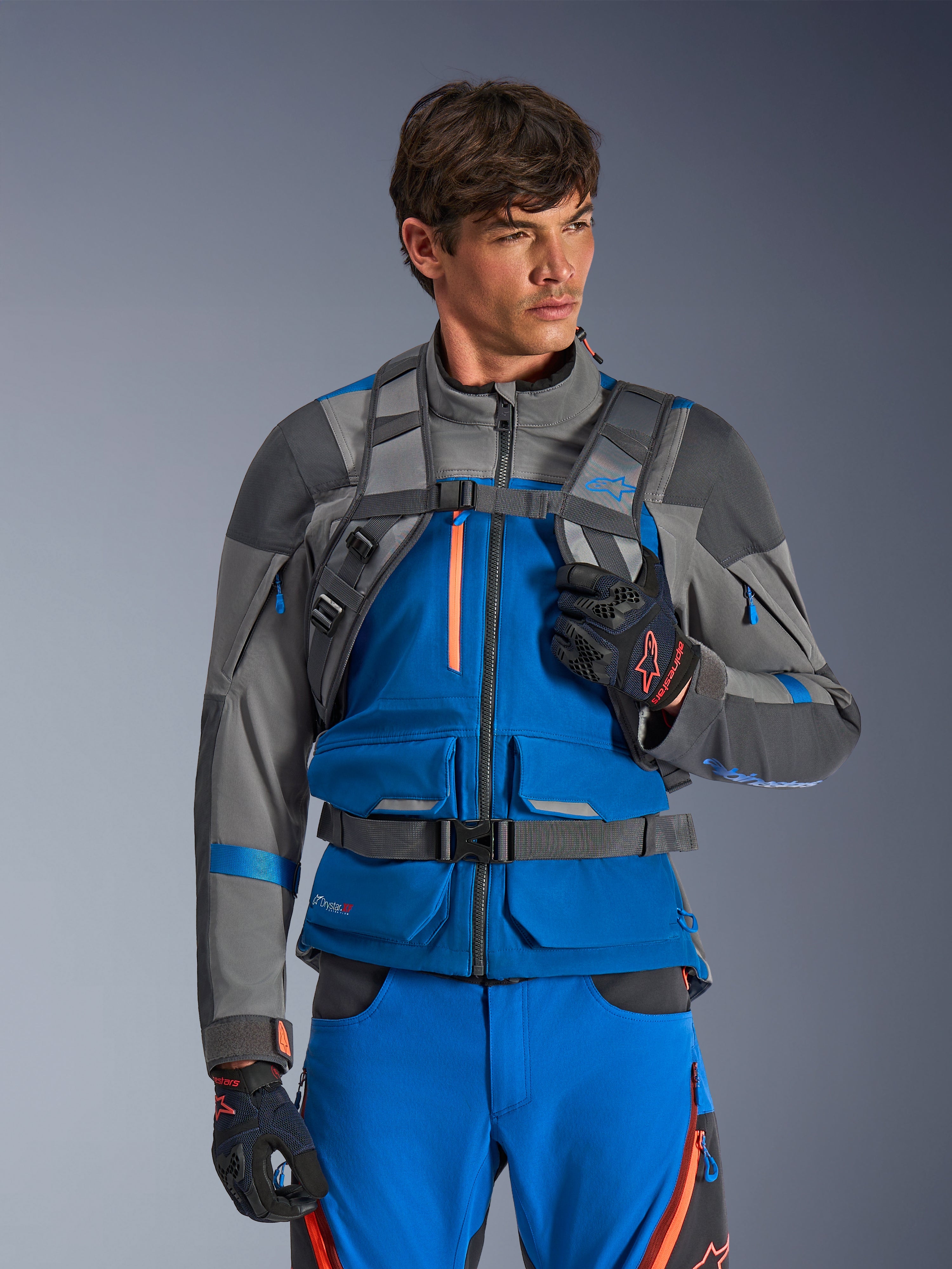 Sac à dos Alpinestars AT-15, sac à dos de moto, gris foncé, porté par un pilote avec une veste et un pantalon d'aventure bleus et gris, doté de bretelles techniques grises avec boucle de poitrine magnétique et sangle ventrale, conception robuste pour la conduite tout-terrain