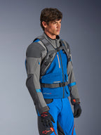 Sac à dos Alpinestars AT-15, sac à dos de moto aventure, gris foncé et gris profond, porté par un pilote sur une veste textile bleue et grise, mettant en évidence le système de harnais technique avec boucles pectorale et ventrale, conçu pour la stabilité en tout-terrain