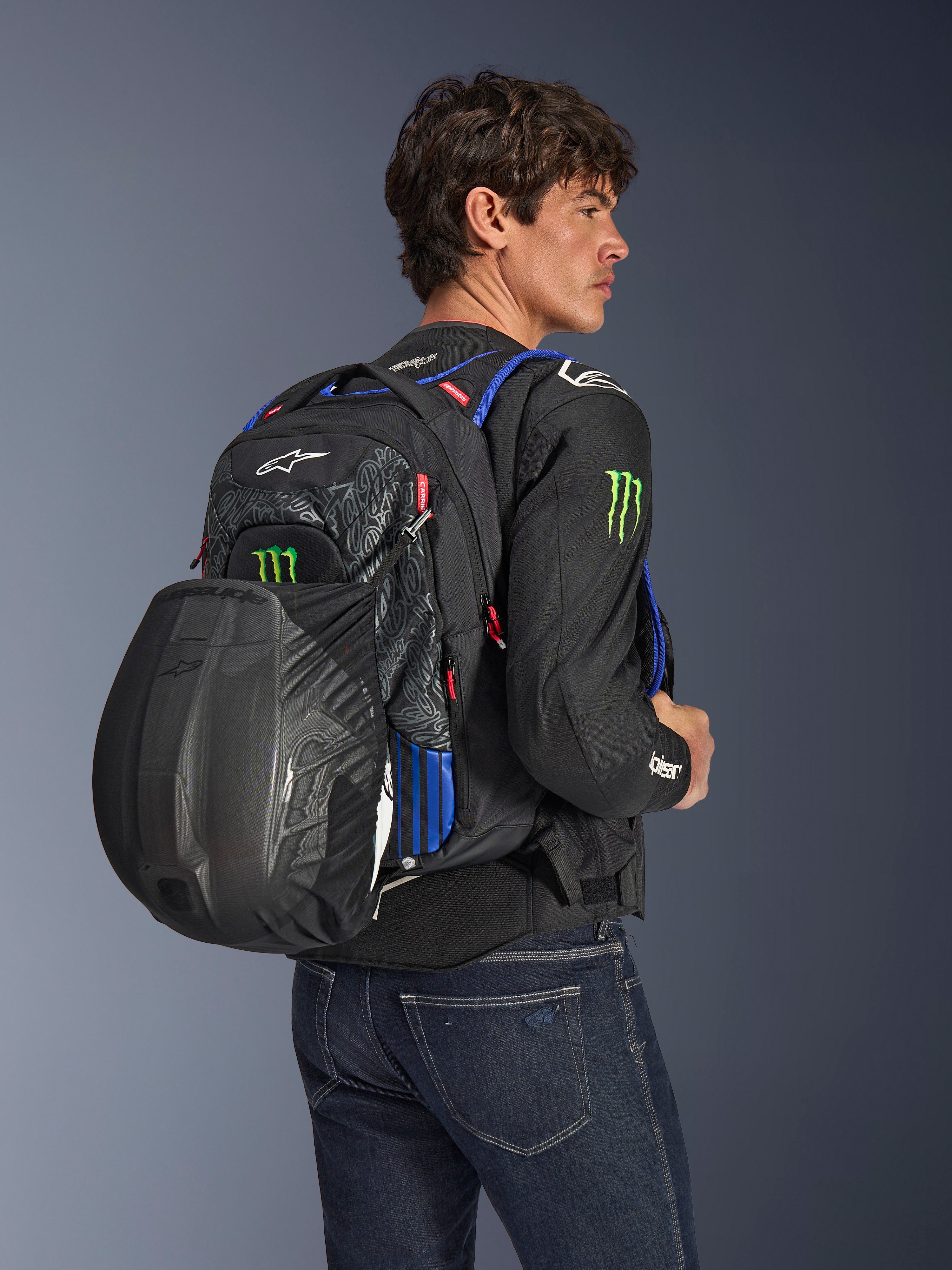 Alpinestars FQ20 City Hunter V2 Monster Backpack, urban motorcycle backpack in Black with Red Fluo, Blue, and Green accents, worn by a model with a black helmet secured in the rear mesh carrier, styled with a black motorcycle jacket and blue jeans.