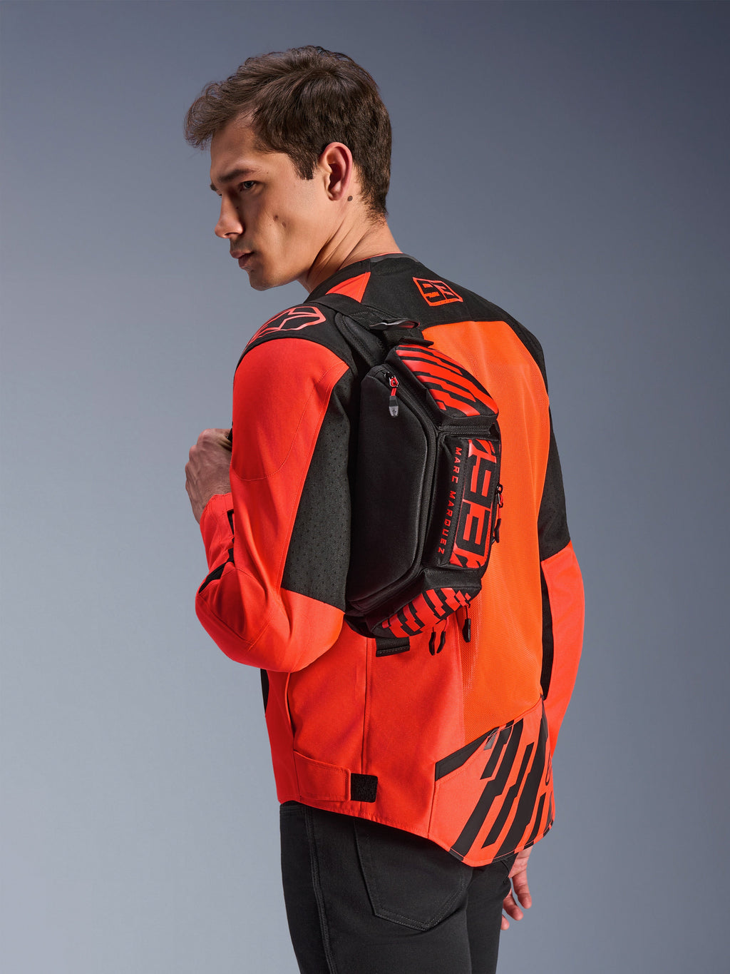 Sac banane Alpinestars MM93, noir avec rouge fluo et rouge vif, porté par un mannequin en bandoulière sur le dos par-dessus une veste de moto rouge et noire, avec logo MM93 et graphismes Marc Marquez