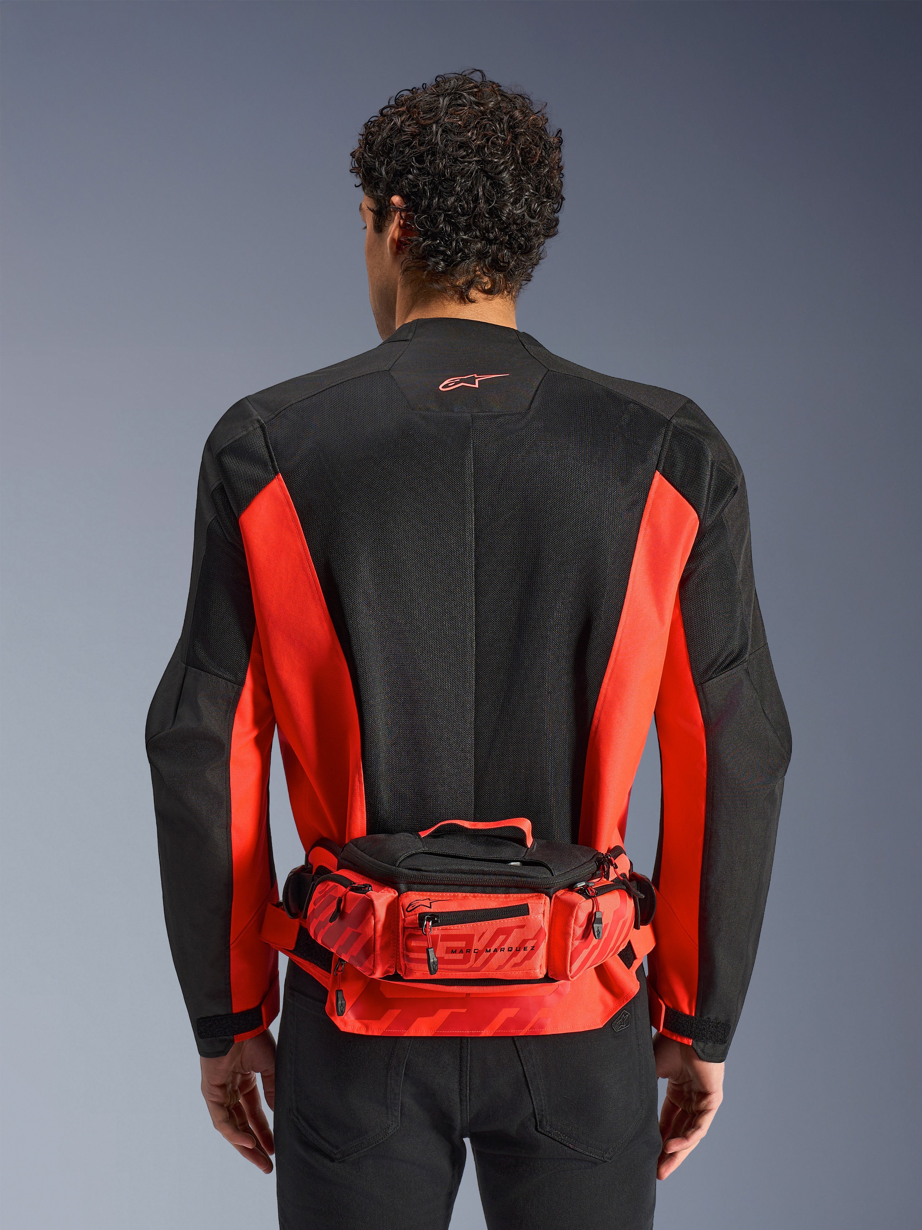 Sac banane Alpinestars MM93, rouge et noir, porté sur un blouson de moto noir et rouge, avec logo Marc Marquez 93, ceinture réglable et compartiments zippés, accessoire fonctionnel d'inspiration racing