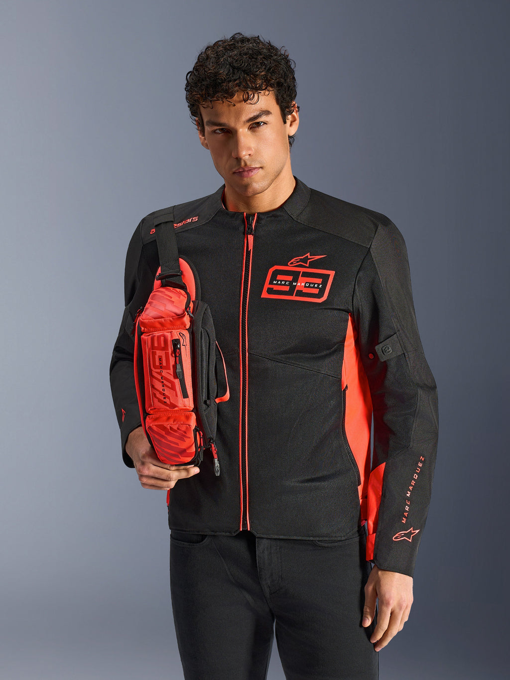 Homme portant le sac banane Alpinestars MM93, rouge fluo et noir, porté en bandoulière sur une veste de moto assortie noire et rouge, avec le logo Marc Marquez 93, design sportif de course