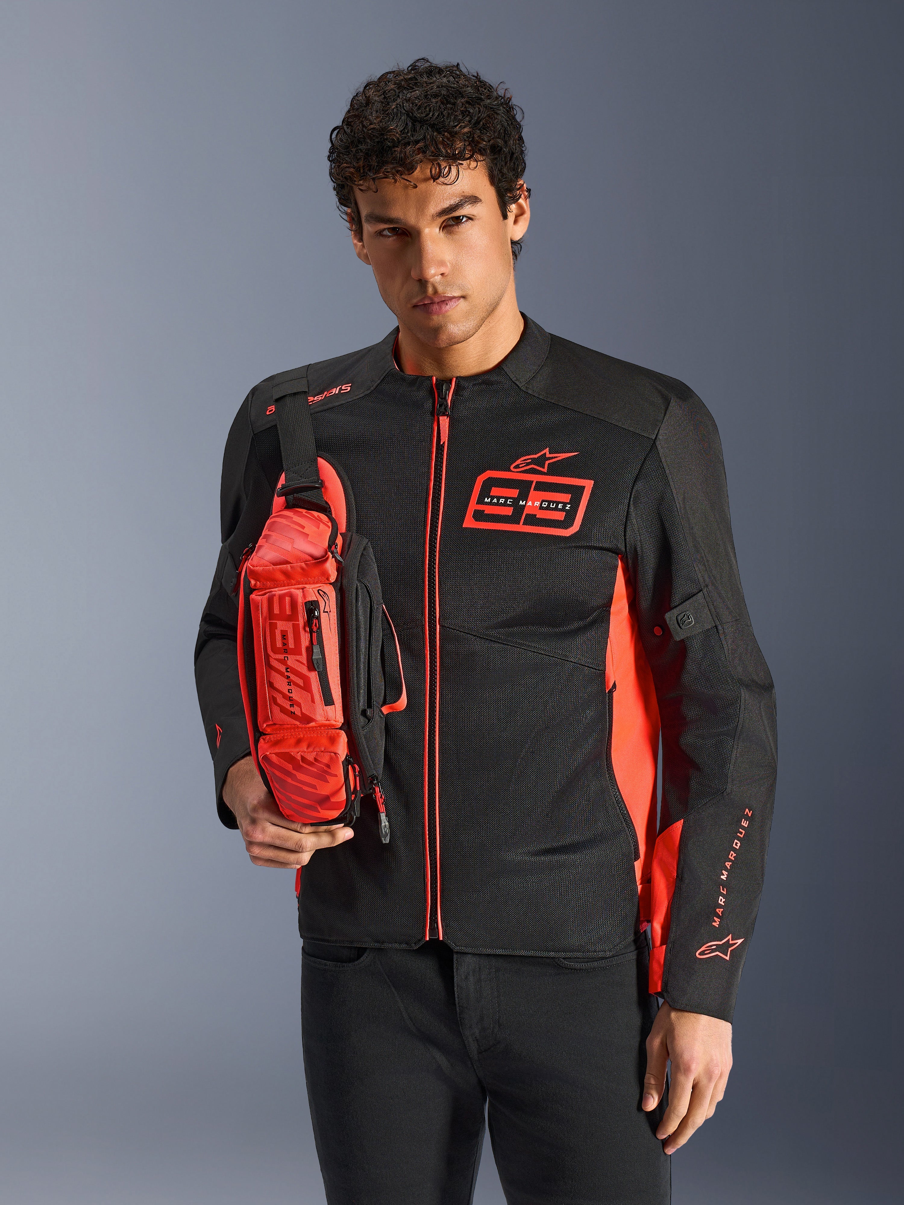 Homme portant le sac banane Alpinestars MM93, rouge fluo et noir, porté en bandoulière sur une veste de moto assortie noire et rouge, avec le logo Marc Marquez 93, design sportif de course