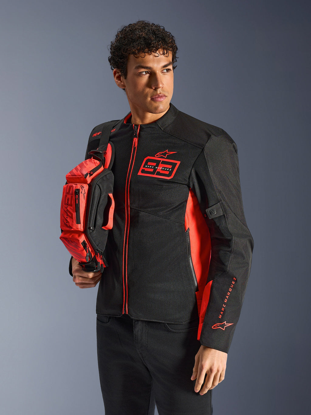 Sac banane Alpinestars MM93, rouge fluo et noir, tenu par un mannequin portant une veste de moto assortie noire et rouge, doté de plusieurs compartiments zippés et du marquage Marc Marquez, équipement pratique pour la conduite sportive