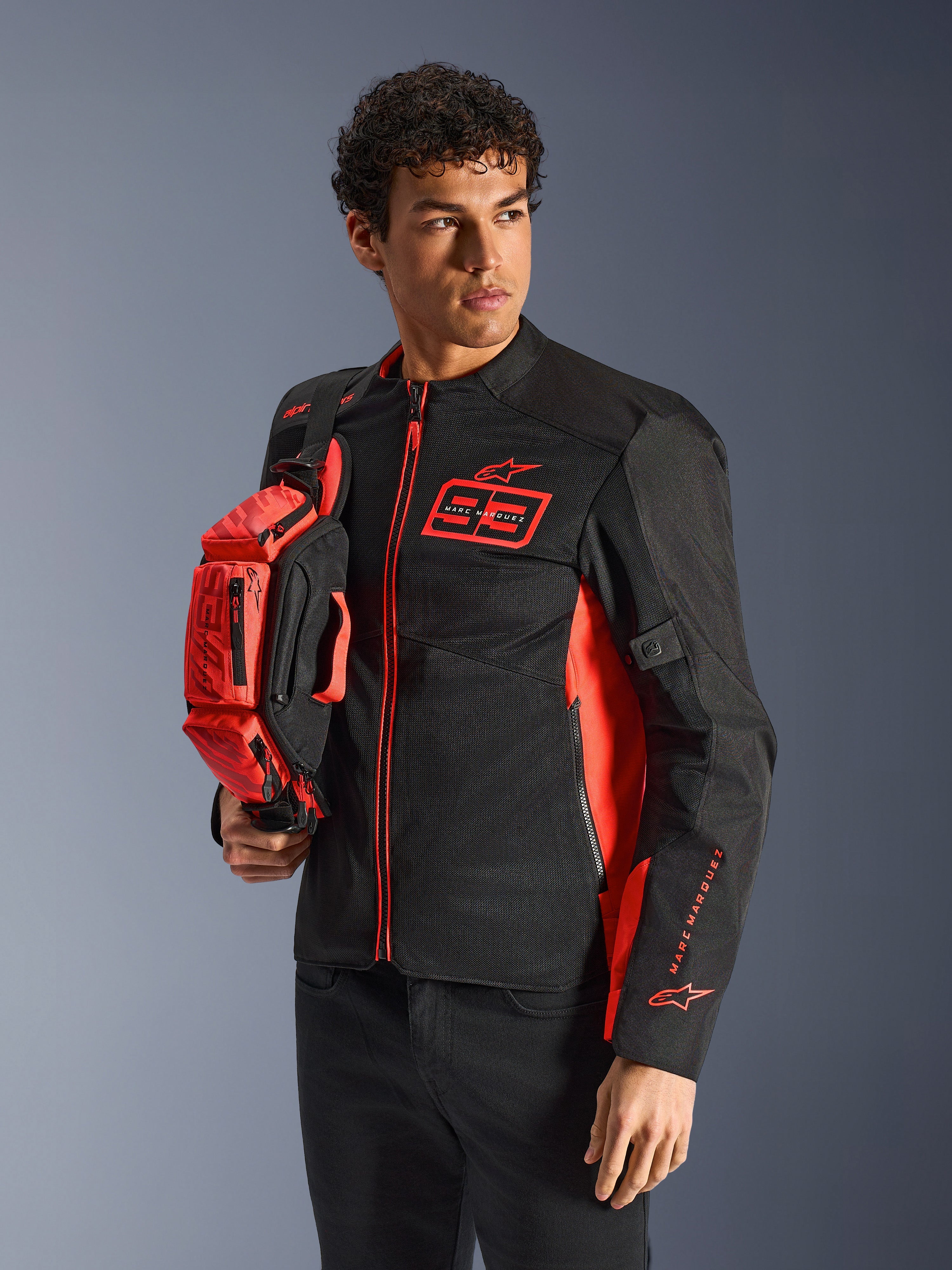 Sac banane Alpinestars MM93, rouge fluo et noir, tenu par un mannequin portant une veste de moto assortie noire et rouge, doté de plusieurs compartiments zippés et du marquage Marc Marquez, équipement pratique pour la conduite sportive