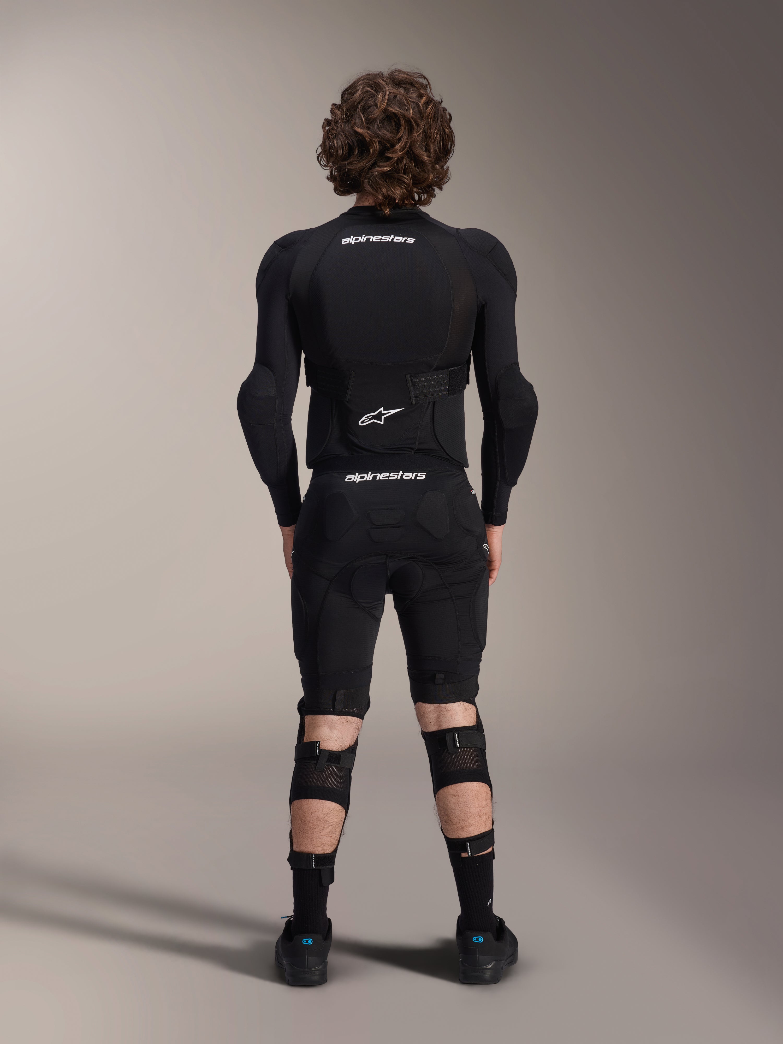 Veste de protection Alpinestars A-Impact Plasma - Manches longues, vue de dos, noire avec logos Alpinestars blancs, portée avec un short de protection et des genouillères pour une protection intégrale en VTT