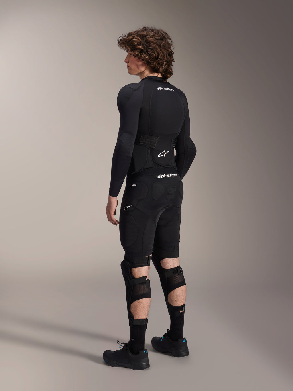 Mannequin portant les protège-genoux et tibias Alpinestars A-Impact Plasma Pro vus de dos, noir et blanc, montrant le système de fermeture à triple sangle et les panneaux en maille ventilée, portés avec un ensemble complet d'équipement de protection de VTT noir comprenant une couche de base à manches longues et un short rembourré