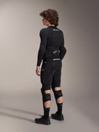 Mannequin portant les protège-genoux et tibias Alpinestars A-Impact Plasma Pro vus de dos, noir et blanc, montrant le système de fermeture à triple sangle et les panneaux en maille ventilée, portés avec un ensemble complet d'équipement de protection de VTT noir comprenant une couche de base à manches longues et un short rembourré