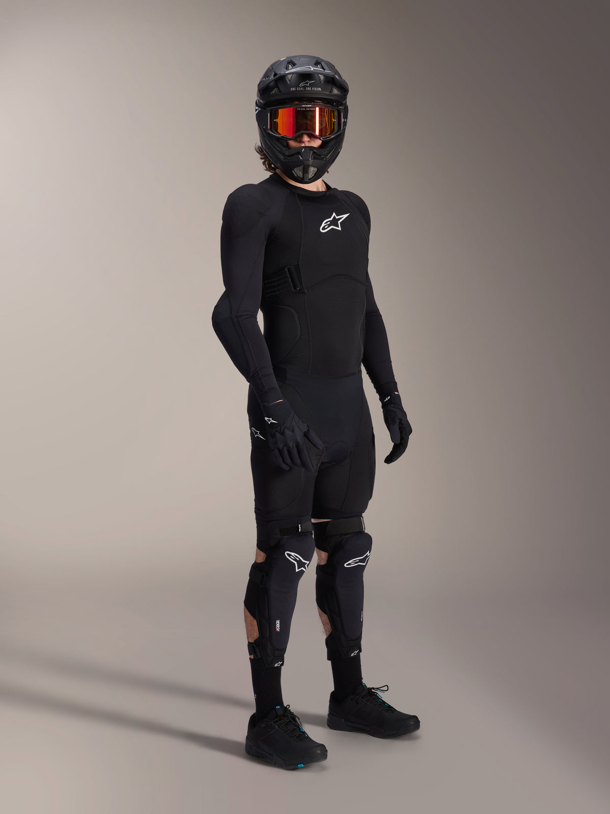 Protections genoux et tibias Alpinestars A-Impact Plasma Pro, noir et blanc, portées par un pilote avec une tenue de protection complète de VTT, incluant un haut blindé noir à manches longues, des gants noirs et un casque intégral avec lunettes orange.