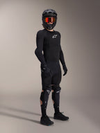 Protections genoux et tibias Alpinestars A-Impact Plasma Pro, noir et blanc, portées par un pilote avec une tenue de protection complète de VTT, incluant un haut blindé noir à manches longues, des gants noirs et un casque intégral avec lunettes orange.