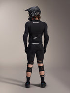 Protège-genoux et tibias Alpinestars A-Impact Plasma Pro, noir, vue arrière sur un rider portant un équipement de protection VTT complet incluant un casque intégral et une protection corporelle, avec système de fermeture à triple sangle et panneaux en maille ventilée pour un maintien optimal et une circulation de l'air