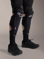 Protections genoux et tibias Alpinestars A-Impact Plasma Pro, noir et blanc, avec logo Alpinestars blanc et système de fermeture à triple sangle, portées avec chaussettes noires et chaussures de sport noires, protection durable pour le cyclisme tout-terrain