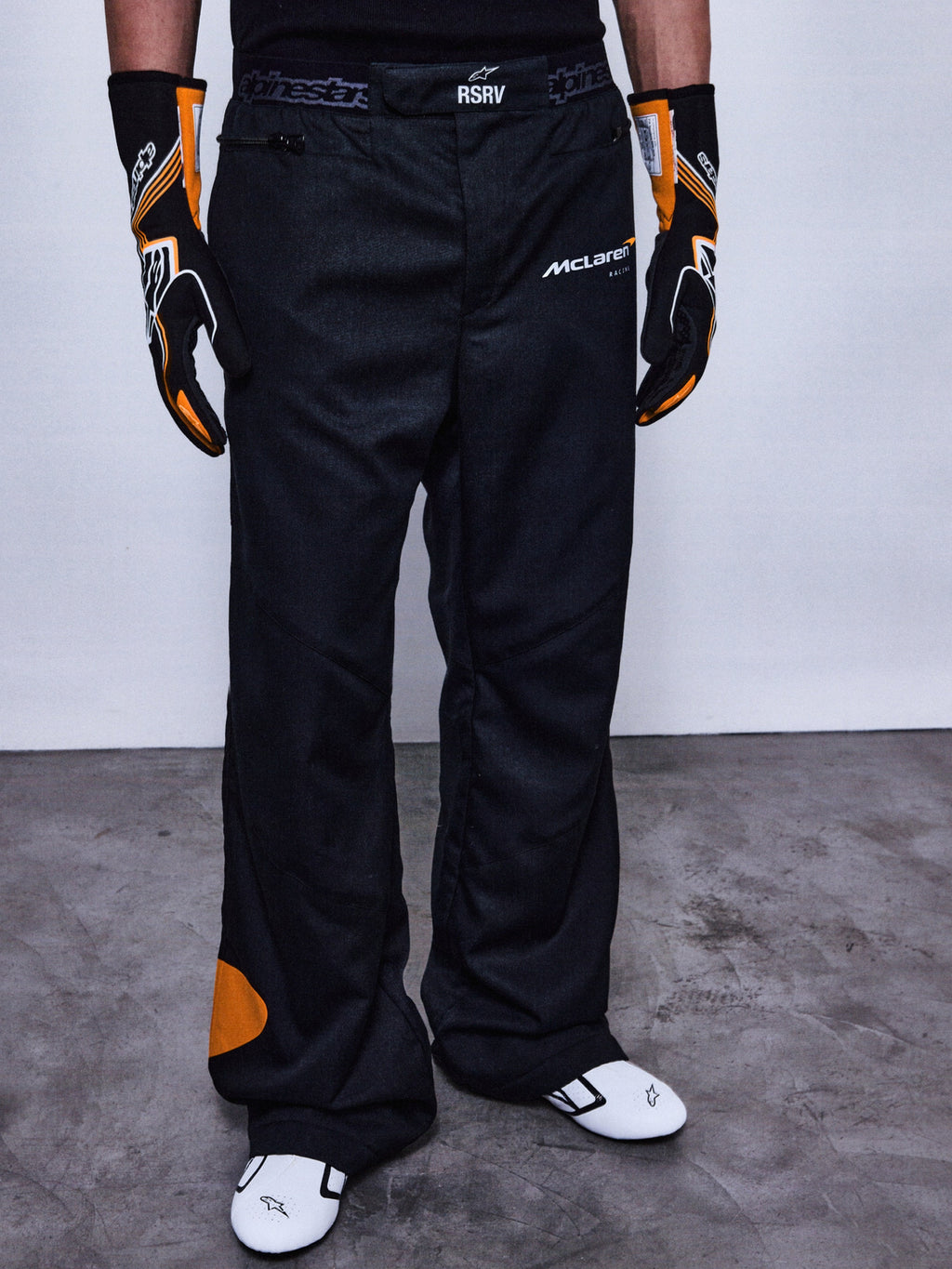 Alpinestars RSRV Arena Suit Pants, pantalon de course noir avec logos McLaren et Alpinestars, détails orange, porté avec gants noirs et orange et chaussures de course blanches