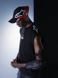 Alpinestars RSRV Tech Tank, débardeur technique sans manches noir avec logo blanc, porté avec coudière de protection texturée noire et casquette rouge et noire