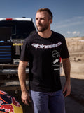 T-shirt Alpinestars Abus CSF, t-shirt à manches courtes, noir, avec un grand logo Alpinestars incurvé et des logos superposés, porté par un homme dans un paddock de sports mécaniques, look lifestyle décontracté