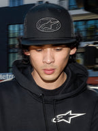 Casquette Trucker Alpinestars Contender, casquette lifestyle noire avec arrière en maille polyester, couronne structurée avec écusson brodé du logo Astars, portée par un homme avec un sweat à capuche Alpinestars noir, streetwear décontracté inspiré du motorsport.