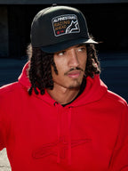 Homme portant une casquette Alpinestars Intuitive Snapback en noir, dotée d'une couronne structurée et d'un écusson brodé « Racing Ahead », associée à un sweat à capuche rouge vif, look streetwear décontracté.