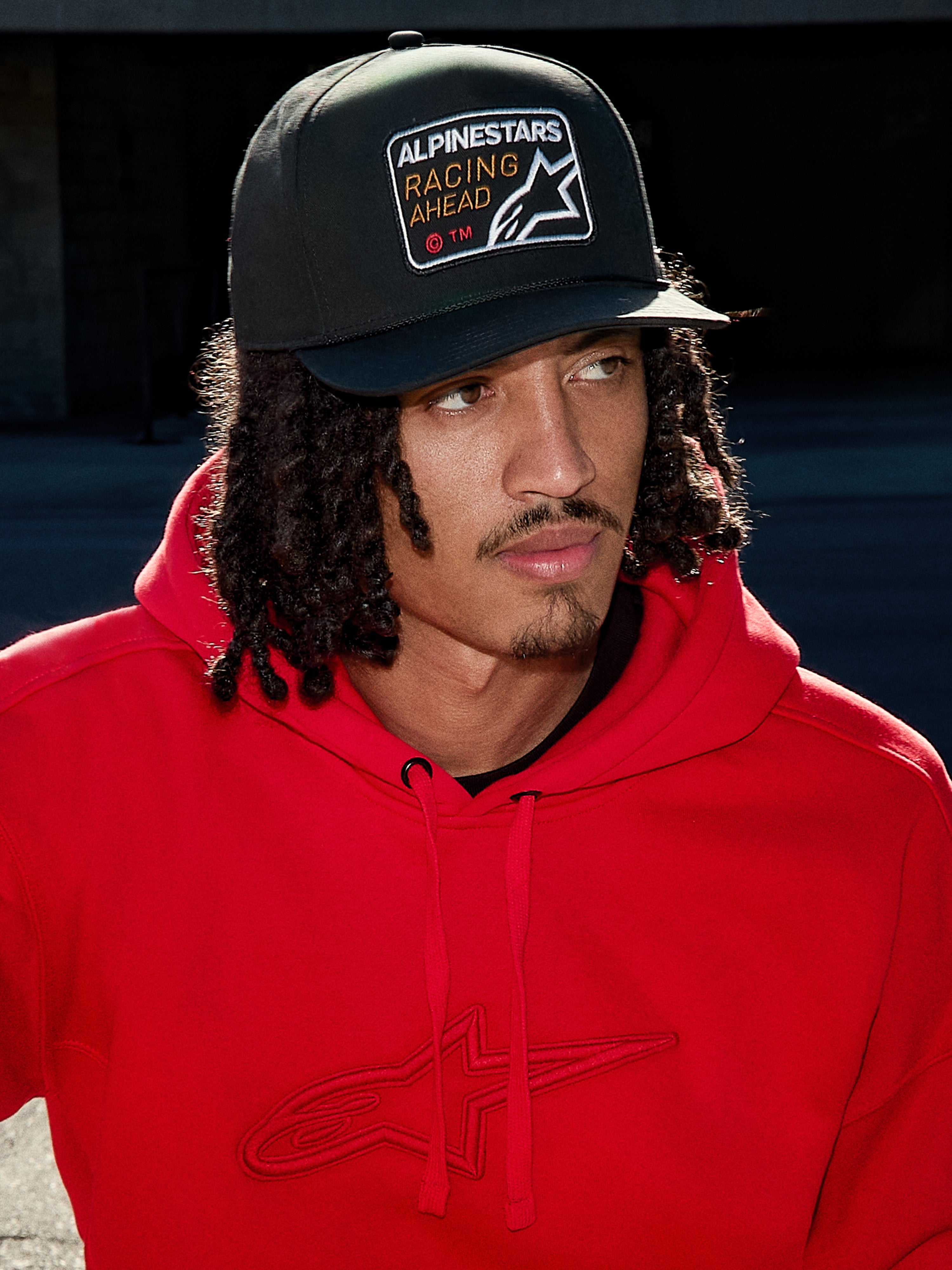 Homme portant une casquette Alpinestars Intuitive Snapback en noir, dotée d'une couronne structurée et d'un écusson brodé « Racing Ahead », associée à un sweat à capuche rouge vif, look streetwear décontracté.