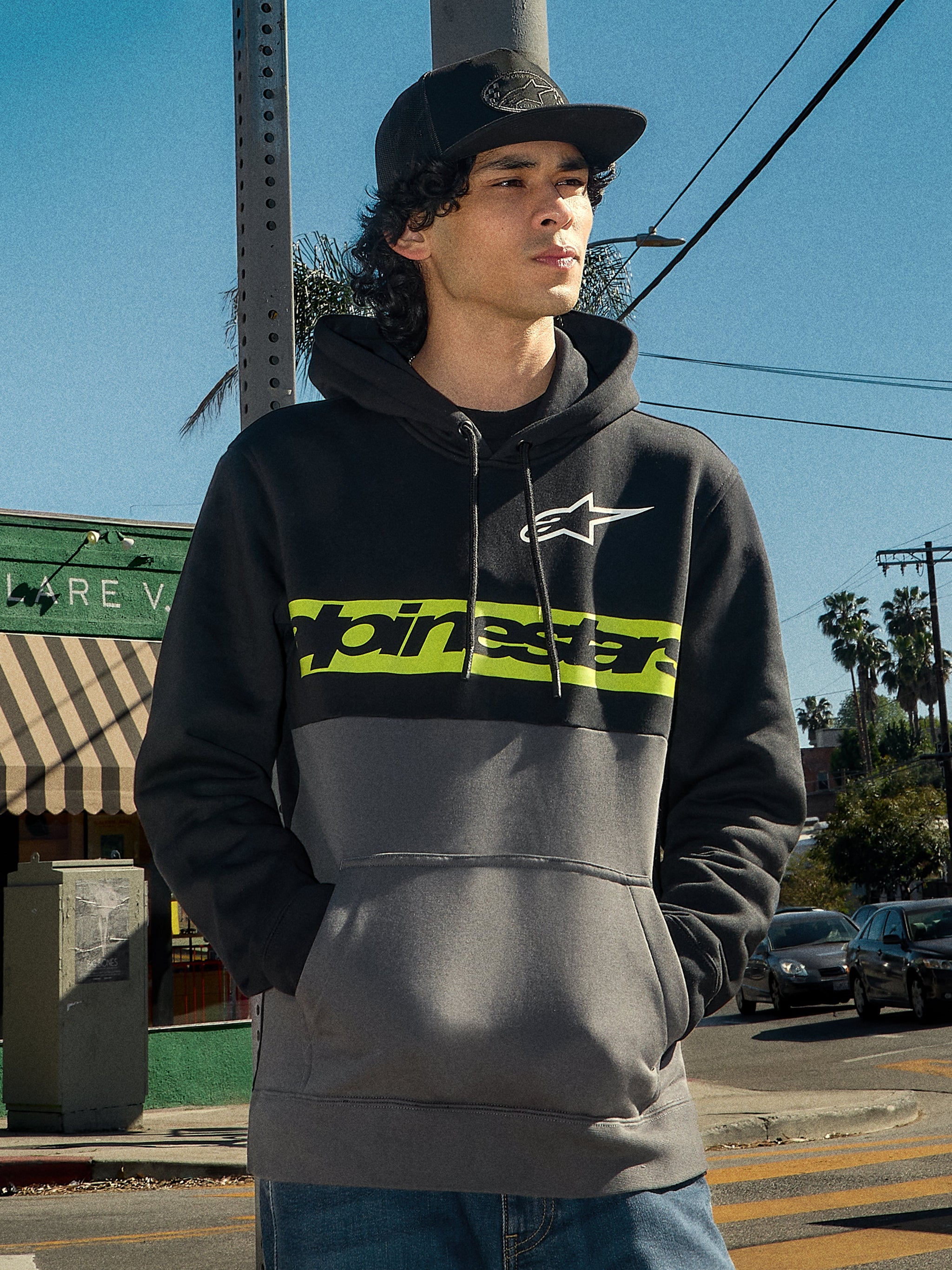 Sweat à capuche Alpinestars Parallel, streetwear lifestyle, design en blocs de couleurs noir et gris anthracite, bande vert citron sur la poitrine avec le logo Alpinestars, logo Astars blanc, capuche à cordon de serrage, porté par un homme dans un environnement urbain extérieur.